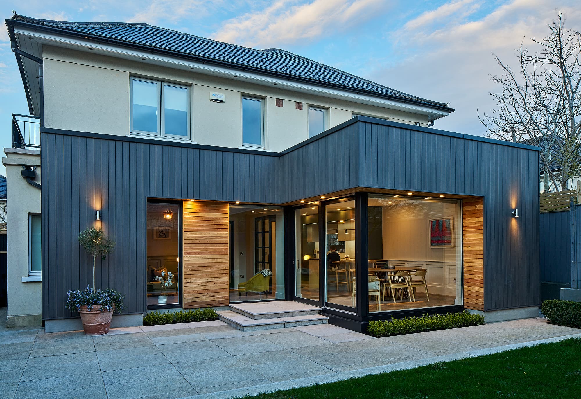 Rear house extension in Carrickmines, Dublin 18 at dusk, grey composite and larch cladding around full-height glazing, by Shomera