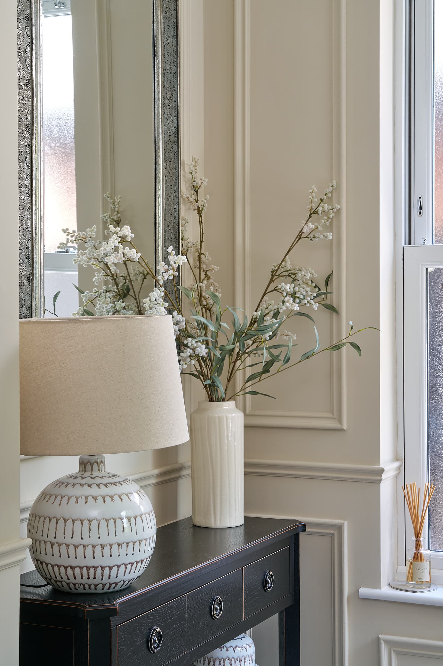 A hallway console table with a ceramic lamp, mirror and vase of blossom in a Shomera house extension