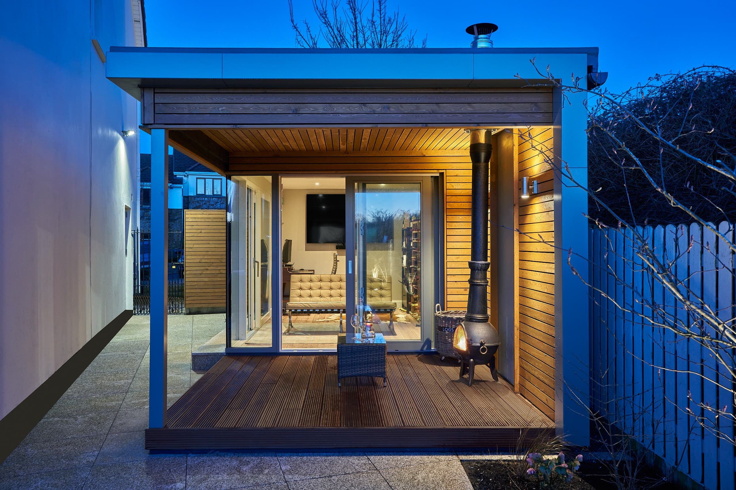 A Shomera garden room in Stocking Wood, Dublin, at dusk, its covered deck and interior lit beneath the overhanging roof