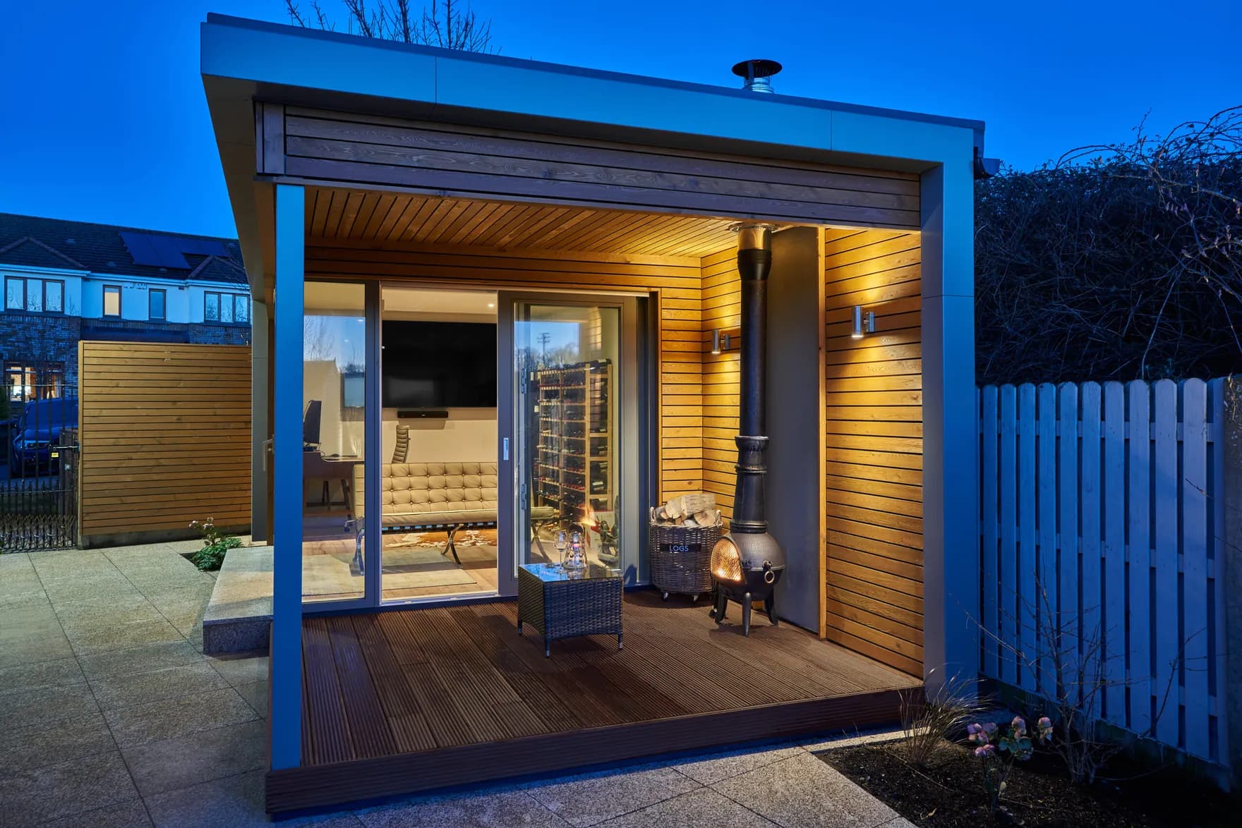 A garden room with a covered timber deck, an outdoor stove and sliding glass doors, at dusk