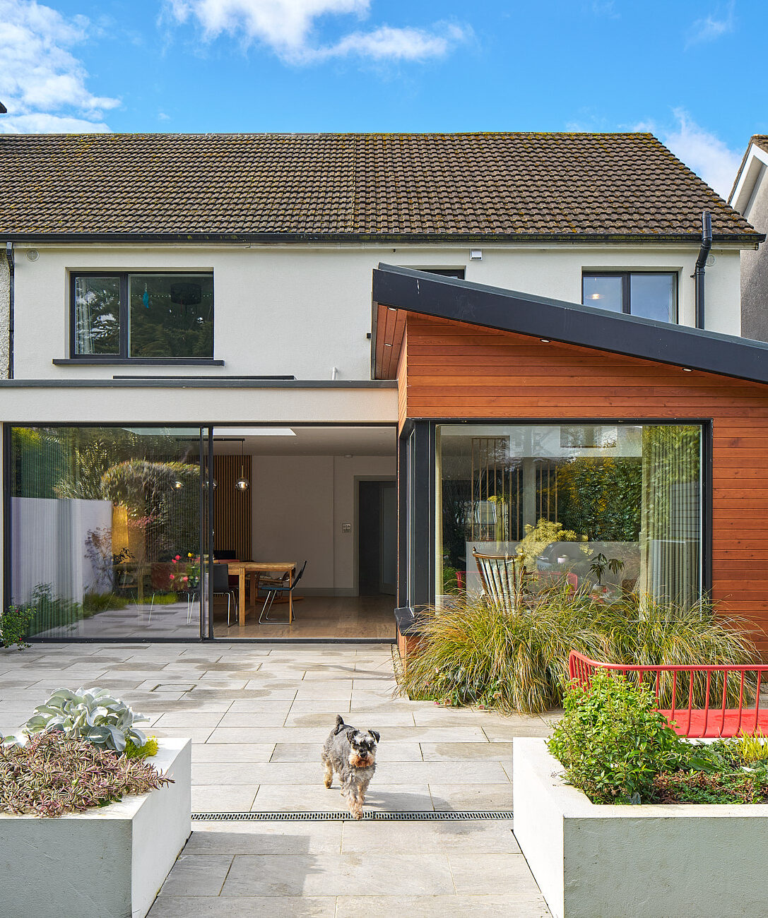 A Shomera house extension with cedar-clad walls and full-height sliding doors opening onto a paved garden terrace, a dog on the path